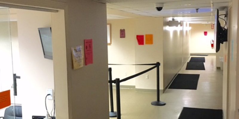 A clean, brightly lit hallway with black floor mats and a roped-off area, leading past a reception window, creating a simple and organized space.