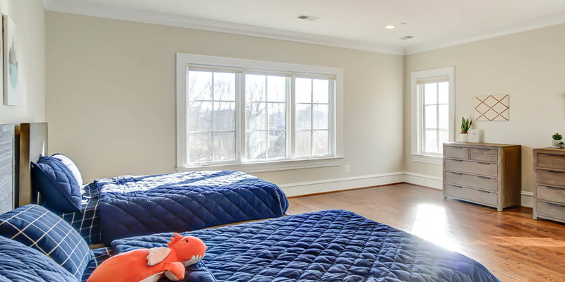 A spacious, sunlit bedroom with two twin beds adorned in blue bedding, cozy wooden floors, and large windows offering a bright, peaceful atmosphere.