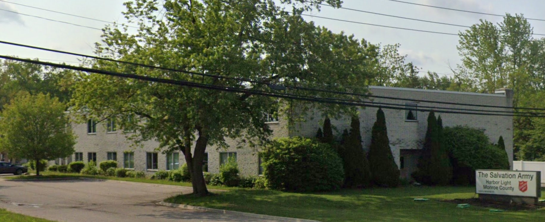 A light-colored building partially hidden by large trees and bushes, with a sign in front.
