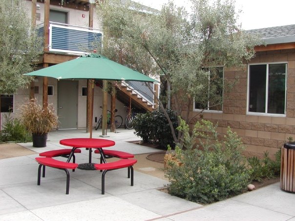 Outdoor courtyard with red picnic table and green umbrella surrounded by trees and shrubs.