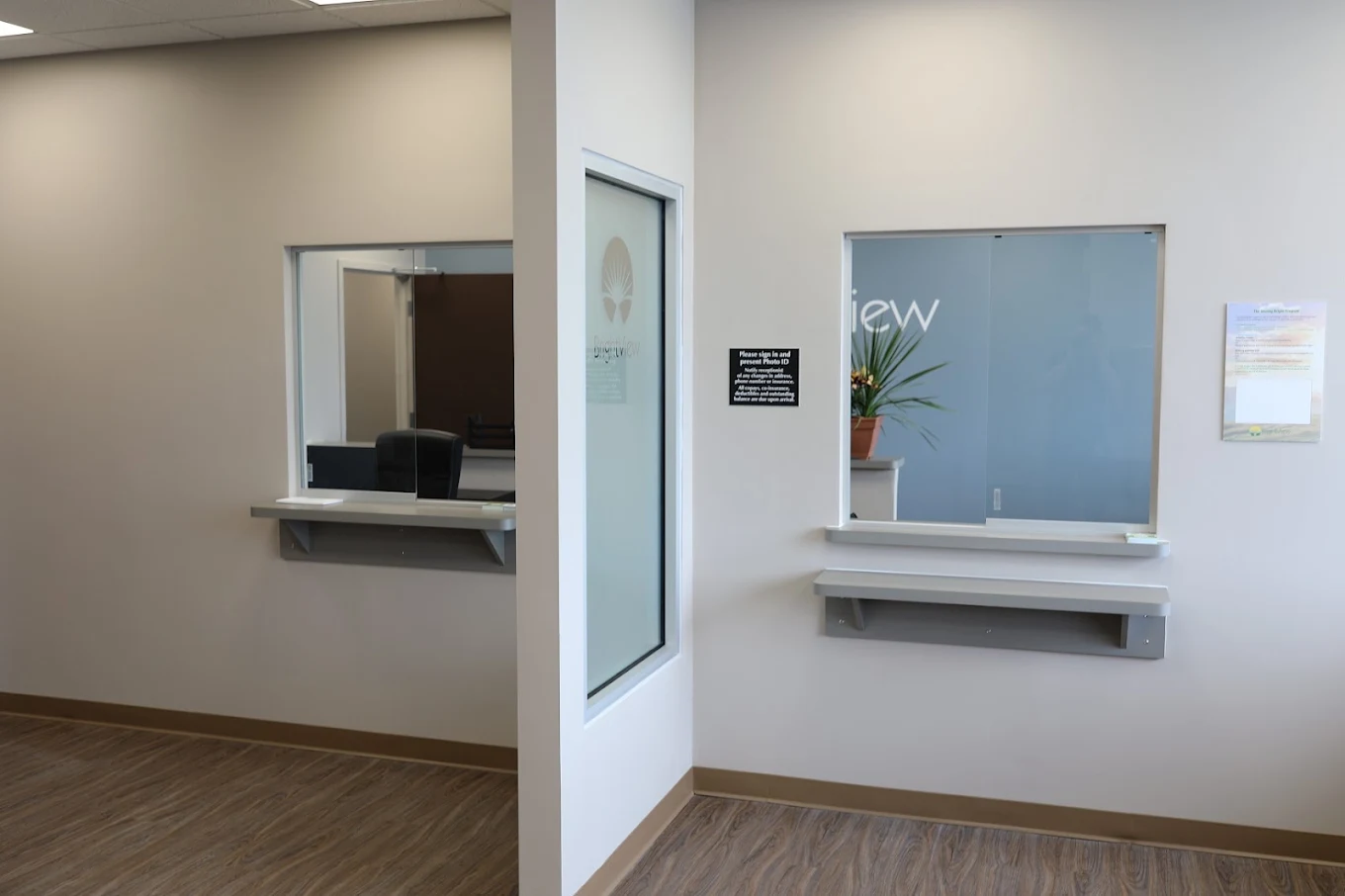 Patient service area with two secure service windows, counter ledges, BrightView-branded glass, and wood-look flooring
