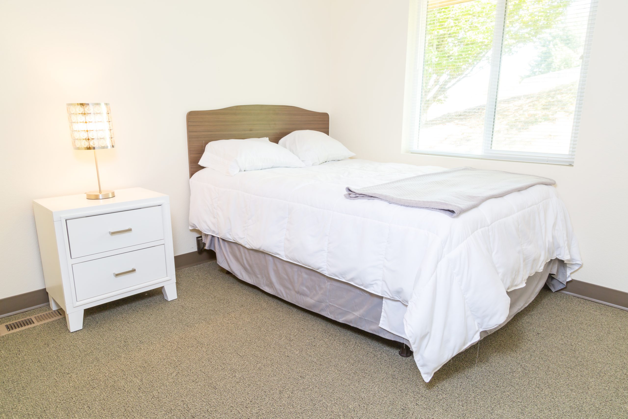 A bedroom with a white bed, nightstand, and window with natural light.