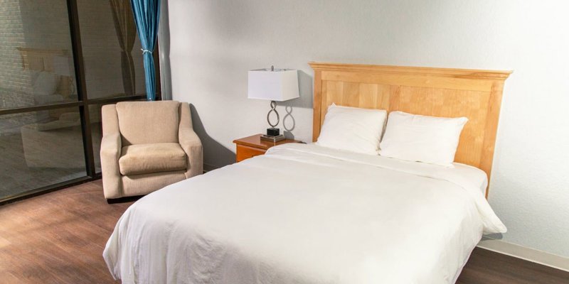 Cozy bedroom with a soft bed, simple wooden headboard, comfortable armchair, and natural light from large windows, offering a peaceful retreat.