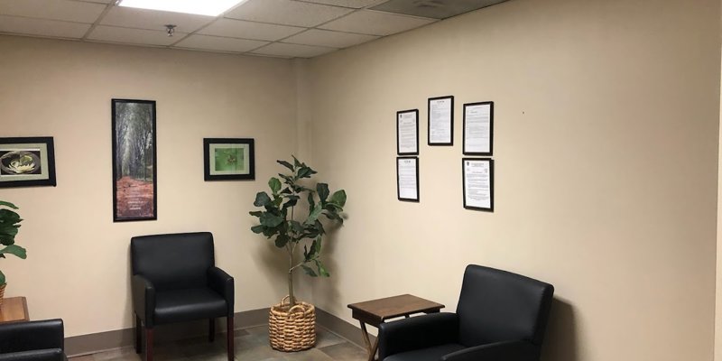 A cozy waiting area with black cushioned chairs, framed artwork, and a potted plant, creating a calm and welcoming atmosphere. Simple yet inviting decor.