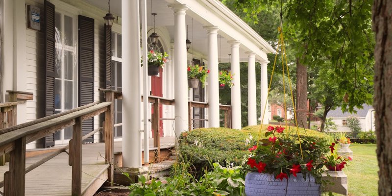 A charming front porch with white columns and hanging flower baskets, surrounded by lush greenery and a peaceful, inviting garden.