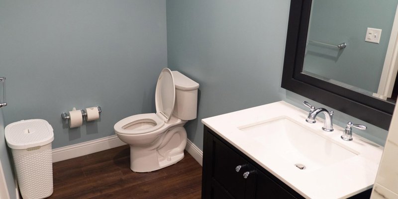 A clean, modern bathroom with light blue walls, a white sink and vanity, dark wood flooring, and a simple wastebasket placed beside the toilet.