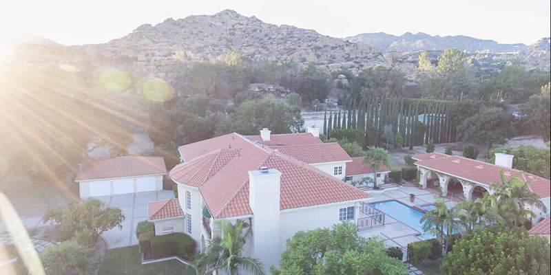 A breathtaking aerial view of a luxurious villa with red-tiled roofs, lush landscaping, a pool, and scenic mountain surroundings illuminated by sunlight.