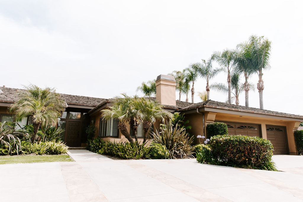 Exterior view of a tan residential-style building with palm trees, lush landscaping, and a wide driveway