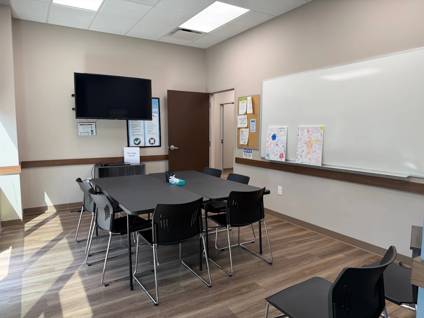 BrightView Health group counseling room with tables and black chairs, a wall-mounted television, large whiteboard, bulletin board, and wood-look flooring