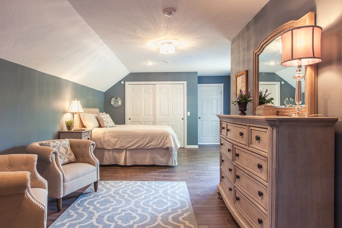 Spacious bedroom with a large bed, armchairs, and a dresser with a mirror in a soft blue color palette