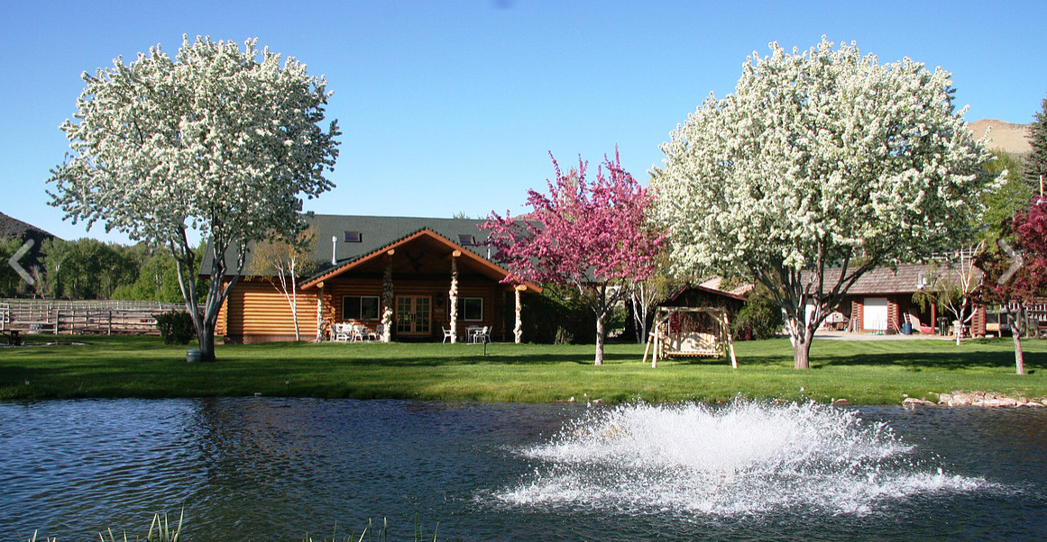 A log-cabin style main lodge framed by blossoming trees and a pond with a water fountain, reflecting the welcoming and well-maintained grounds of Rainbows End Recovery Center in Challis