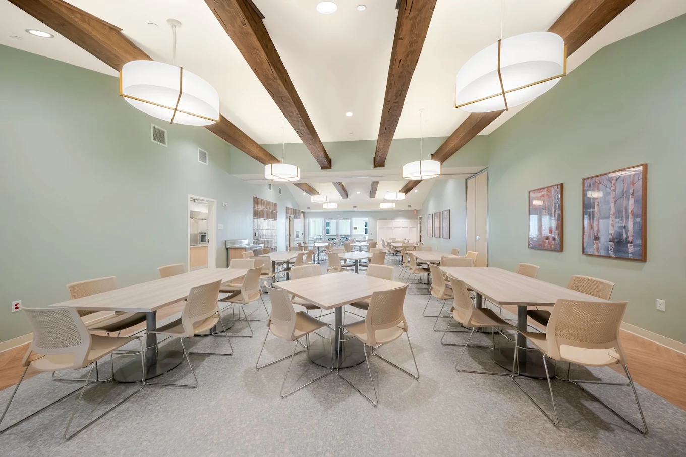 Bright dining and common area with tables, chairs, exposed wood ceiling beams, and large windows
