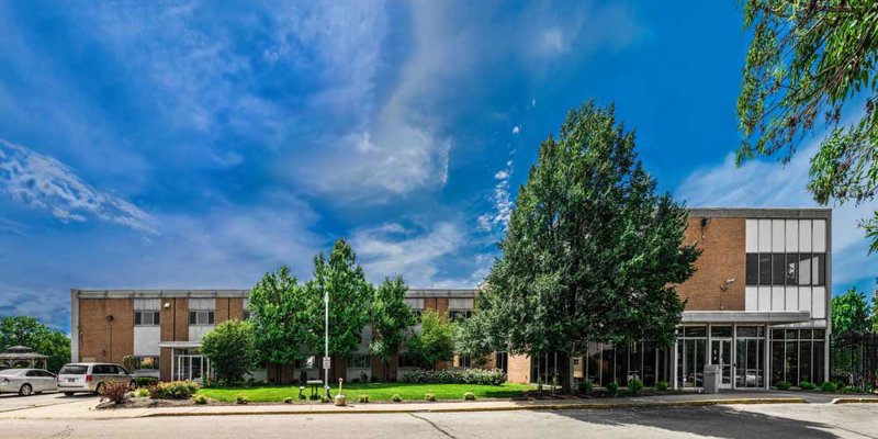 A modern facility with large windows, greenery, and spacious parking, providing a welcoming atmosphere under a bright blue sky.