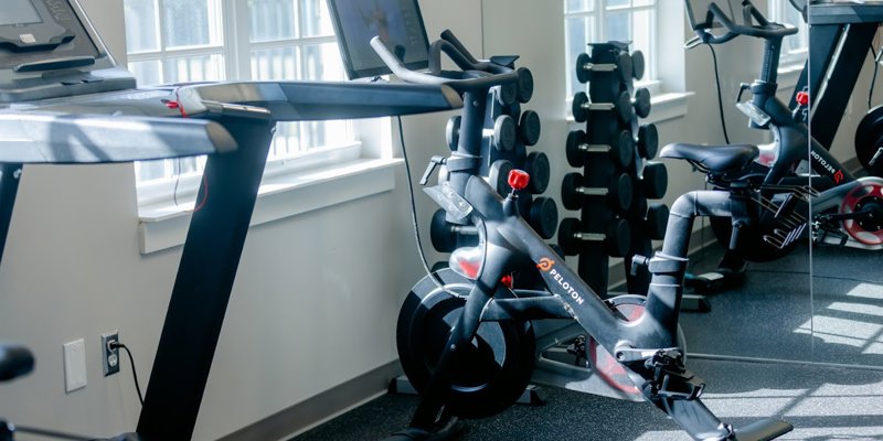 A bright fitness area featuring a treadmill, Peloton bike, and a set of dumbbells. The space is filled with natural light from large windows.