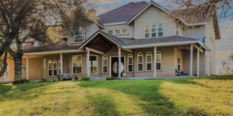 A beautiful two-story home with a large, welcoming porch, surrounded by lush greenery and tall trees, basking in warm sunlight.