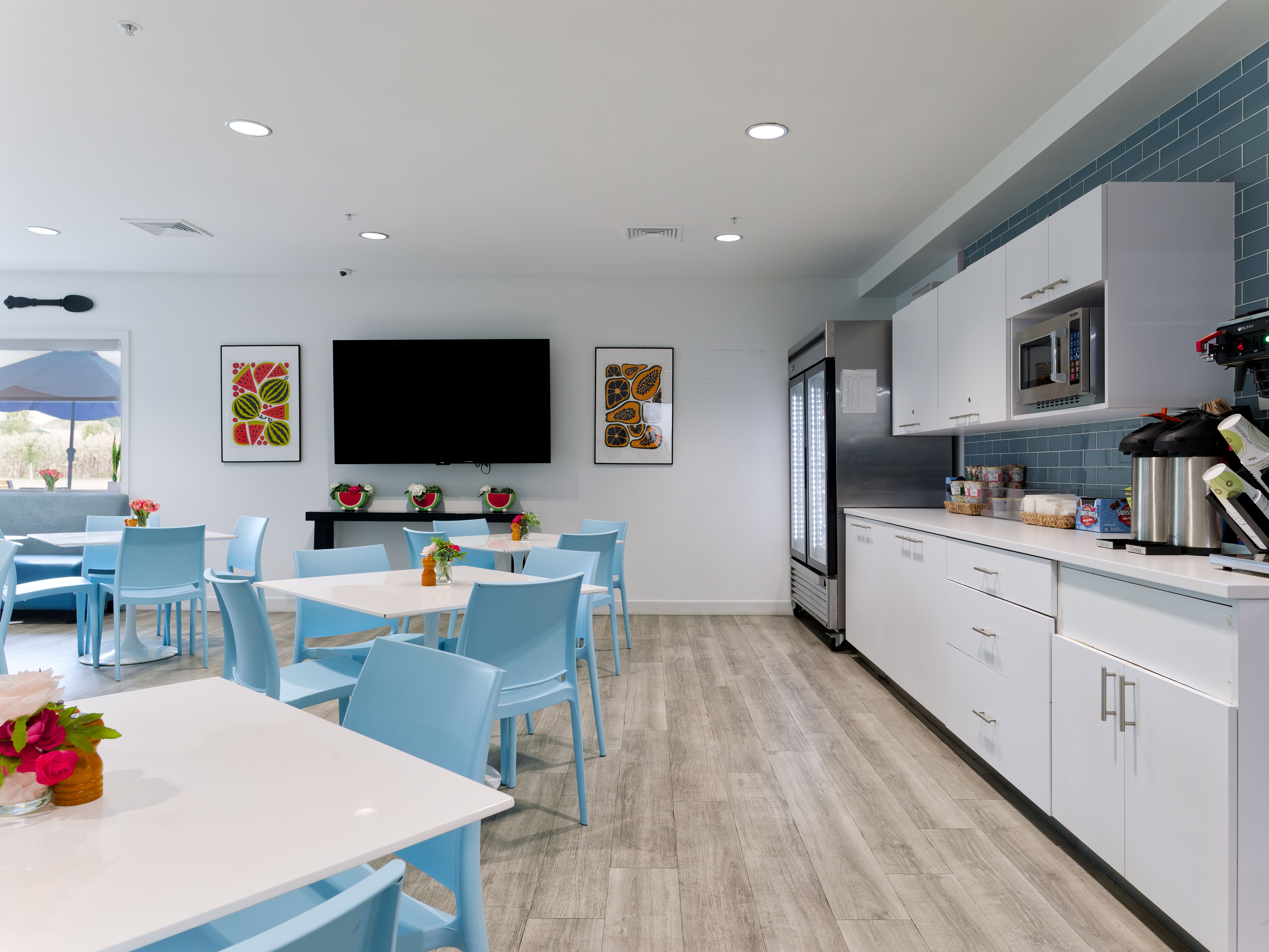 Bright cafeteria with blue chairs, white tables, and TV.