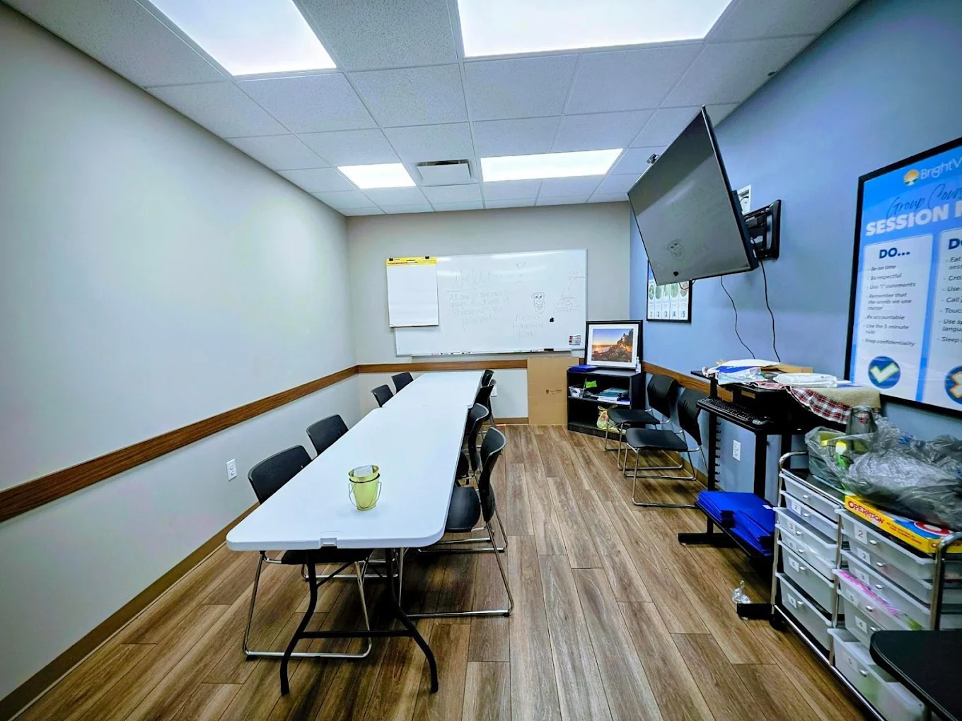 Group counseling room at BrightView Health with long table, chairs, whiteboard, wall-mounted television, storage shelves, and blue accent wall