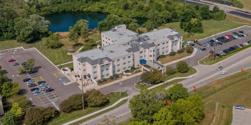 A serene aerial view of a large white building nestled by a tranquil lake, surrounded by lush greenery and ample parking space nearby.