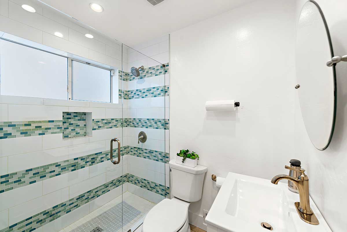 Bright bathroom with white vanity, toilet, round mirror, and glass-enclosed shower with blue and green mosaic tile accents