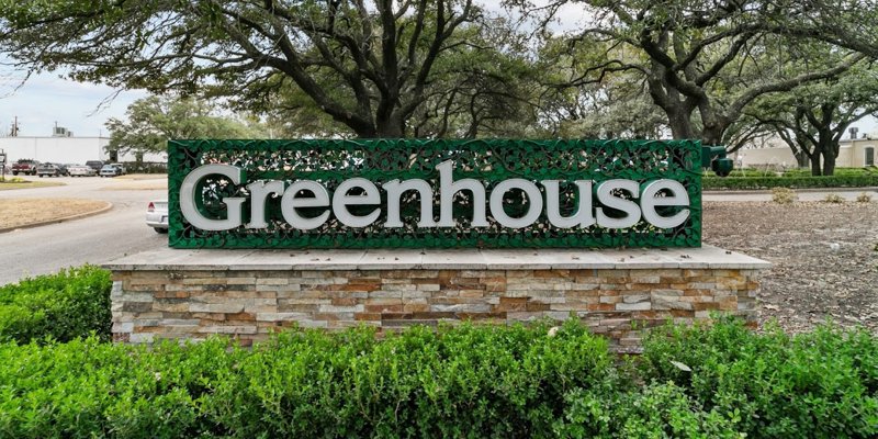  Bold "Greenhouse" sign with vibrant green accents and leafy details, set against a brick base and surrounded by lush greenery.