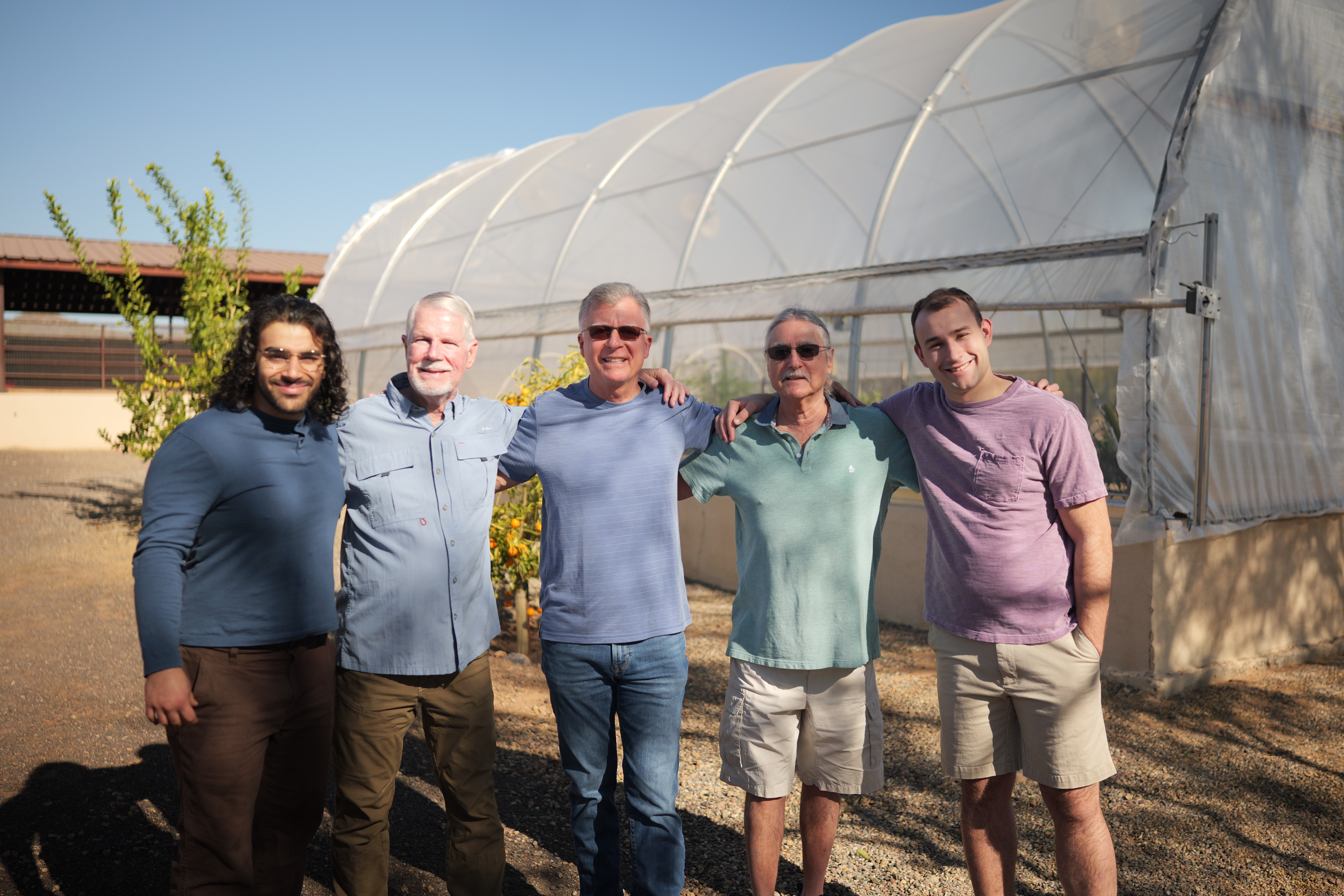 Five men smiling together outside near a greenhouse