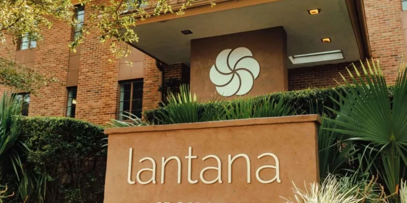 Warm and inviting entrance to Lantana at 65 Gadsden, surrounded by greenery and featuring a modern sign with a distinctive logo on a brick building.