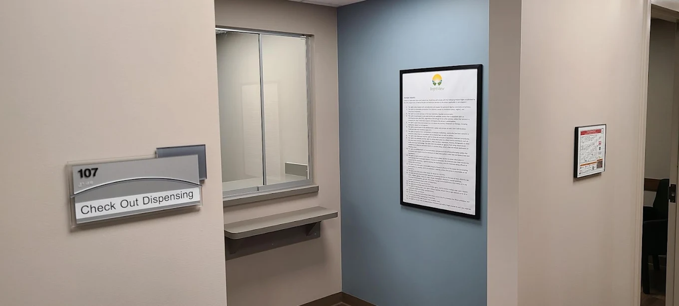 BrightView Health check-out dispensing area with service window and counter, room 107 sign, blue accent wall, and framed clinic information