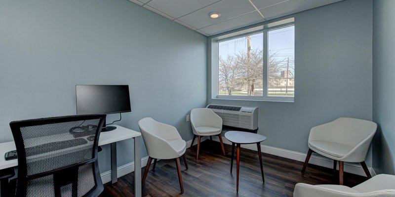 A private, comfortable office space with modern furnishings, featuring a desk with a computer, white chairs, and a window providing natural light.