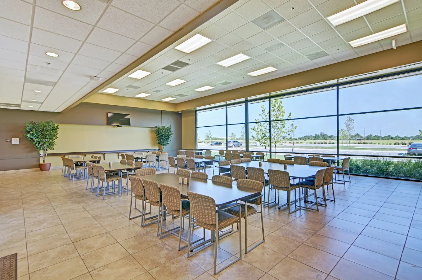 A large dining area with multiple tables and chairs, floor-to-ceiling windows providing natural light, and a television mounted on the wall.