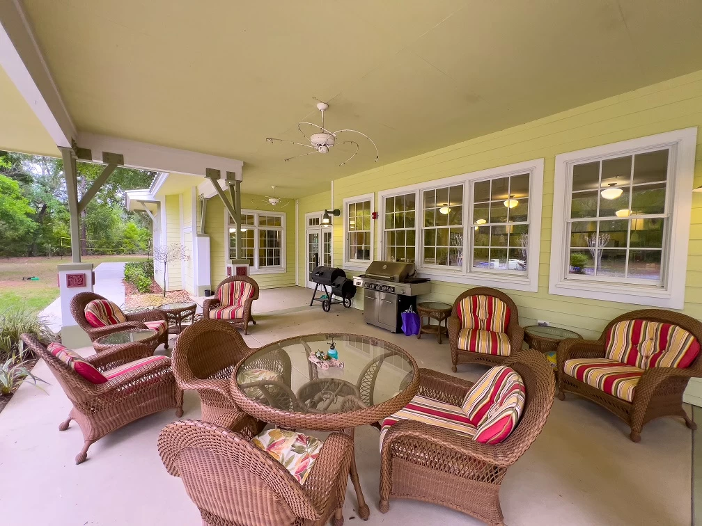 Covered outdoor patio with wicker furniture, dining table, and grill area surrounded by nature