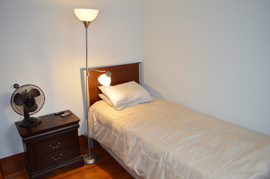 Single bed with beige comforter next to a nightstand, lamp, and small fan