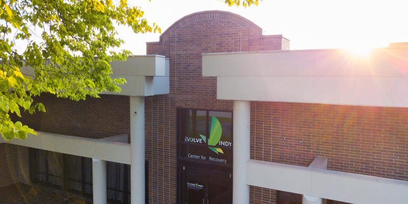 Sunlit entrance of a recovery center with a brick facade, large windows, and lush green leaves, creating a warm and welcoming atmosphere.