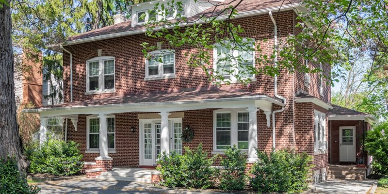 A charming brick building with white trim and a welcoming front porch surrounded by lush greenery and mature trees, creating a peaceful and homey atmosphere.