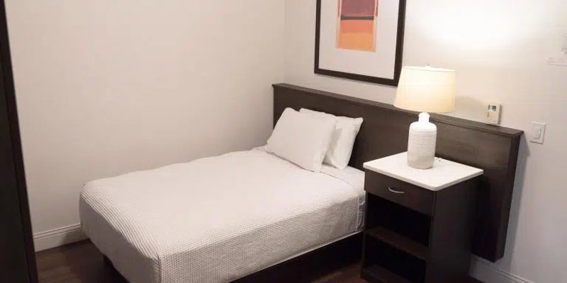 A cozy, minimalist bedroom with a neatly made bed, a bedside table with a lamp, wooden flooring, and warm artwork hanging above the headboard.