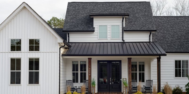 A charming white house with a black metal roof, large windows, and a cozy front porch featuring rocking chairs and neatly trimmed plants.