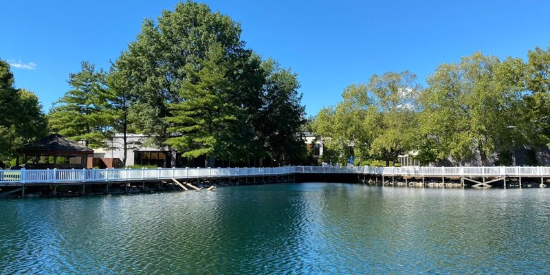 Peaceful lakeside view with a boardwalk surrounded by lush trees, a charming gazebo, and clear blue skies.