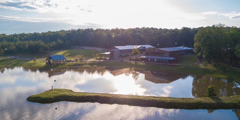 A serene lakeside setting with modern buildings surrounded by lush greenery, reflecting on calm waters under a bright, clear sky.