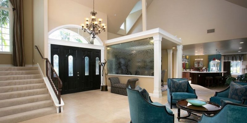 A grand and elegant living space featuring high ceilings, a modern chandelier, plush teal seating, and a beautiful staircase near the entryway.