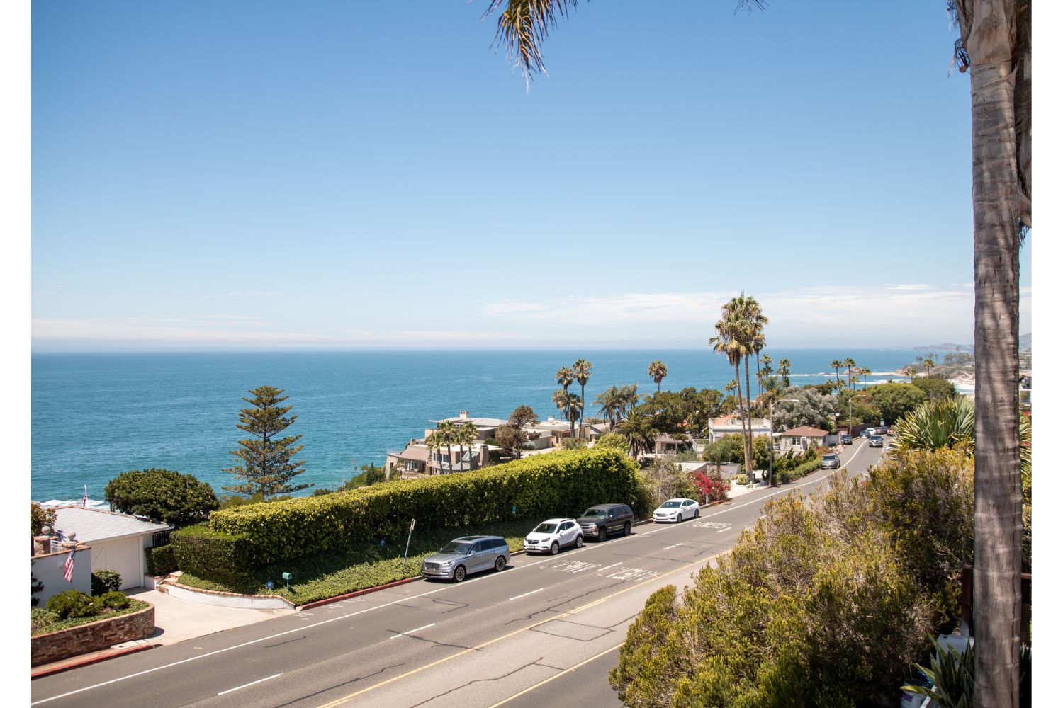 Scenic coastal view featuring palm trees, a blue ocean, and a sunlit road with cars driving along, highlighting a peaceful seaside atmosphere.