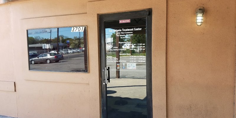 The entrance of a treatment center features a glass door displaying operating hours, with a beige exterior wall and a window reflecting the street.