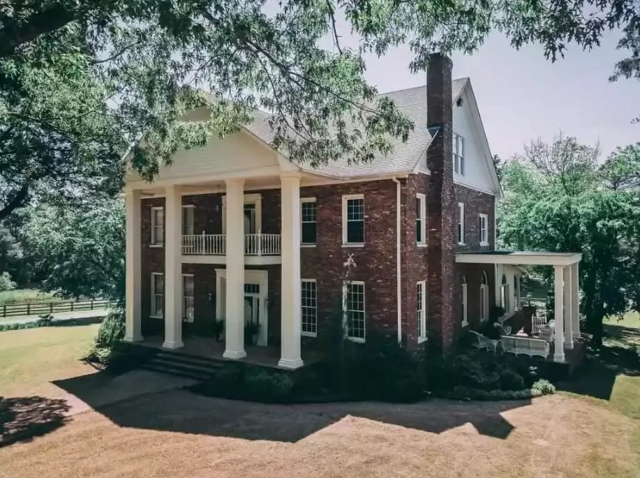 Large two-story brick residence with tall white columns, covered porches, mature trees, and landscaped grounds