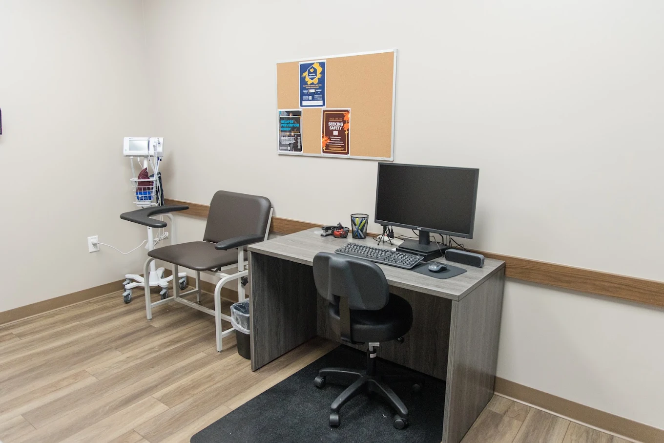 Clinical room with patient chair, vital-sign monitor, computer workstation, bulletin board, and staff chair