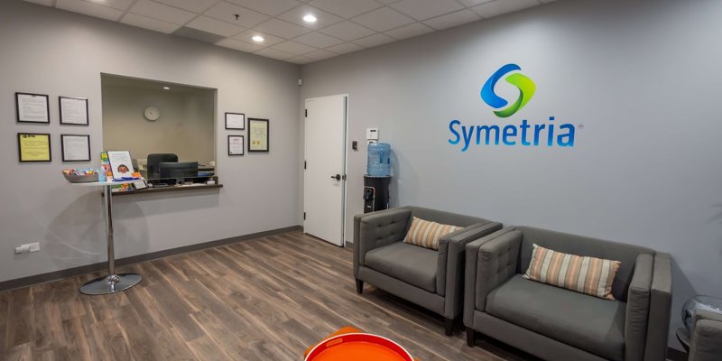 A modern waiting room with comfortable gray chairs, a water cooler, and the Symetria logo on the wall, offering a clean and inviting atmosphere for visitors.