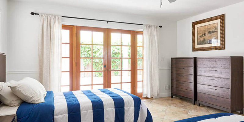 A bright, airy bedroom with twin beds adorned in blue and white bedding, wooden dressers, and large glass doors that invite natural light.