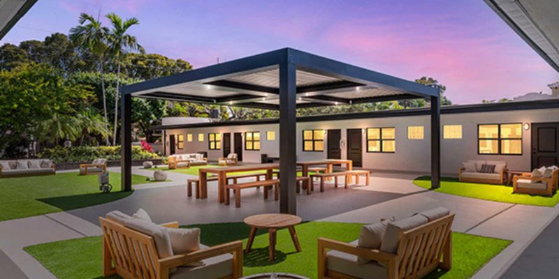 A beautifully lit outdoor courtyard at dusk with comfortable seating, a covered picnic area, and lush greenery, creating a serene and inviting atmosphere.