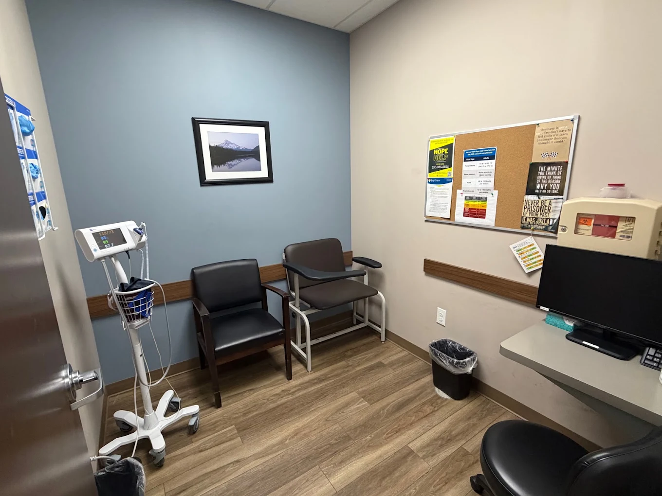 BrightView Health clinical room with patient chairs, vital signs monitor, computer workstation, bulletin board, and blue accent wall