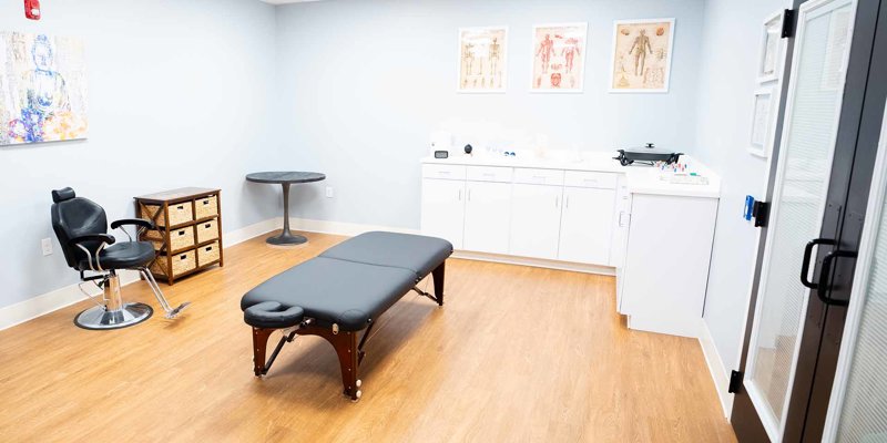 Clean and well-organized therapy room with a massage table, comfortable chair, and bright, welcoming atmosphere designed for relaxation.