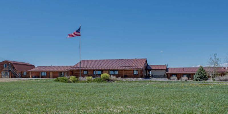 A spacious ranch-style building with a red roof, an American flag proudly waving, surrounded by green fields and set against a clear blue sky.