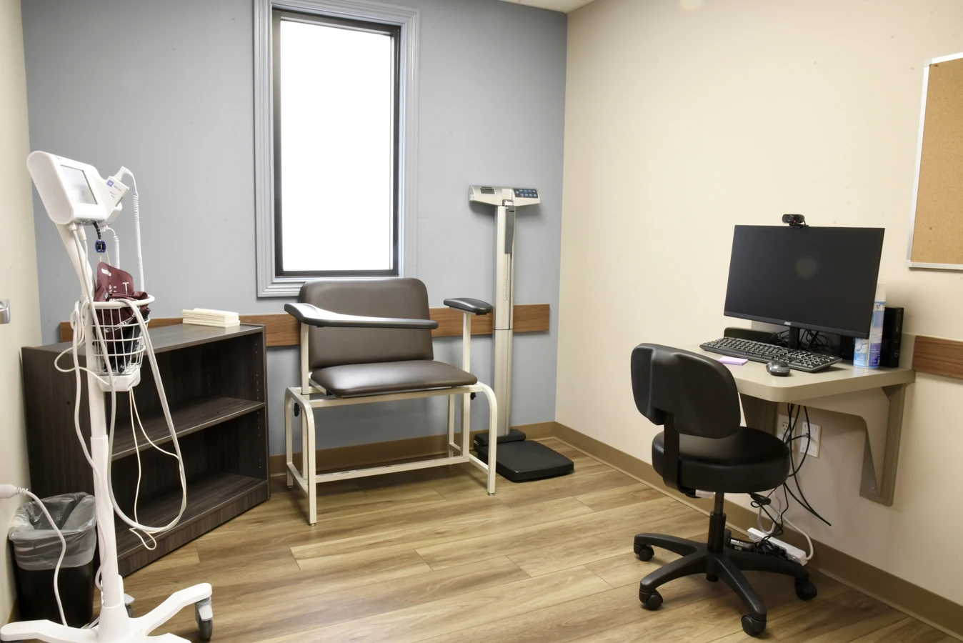 BrightView Health clinical exam room with medical monitoring equipment, bariatric exam chair, standing scale, computer workstation, rolling chair, and wood-look flooring