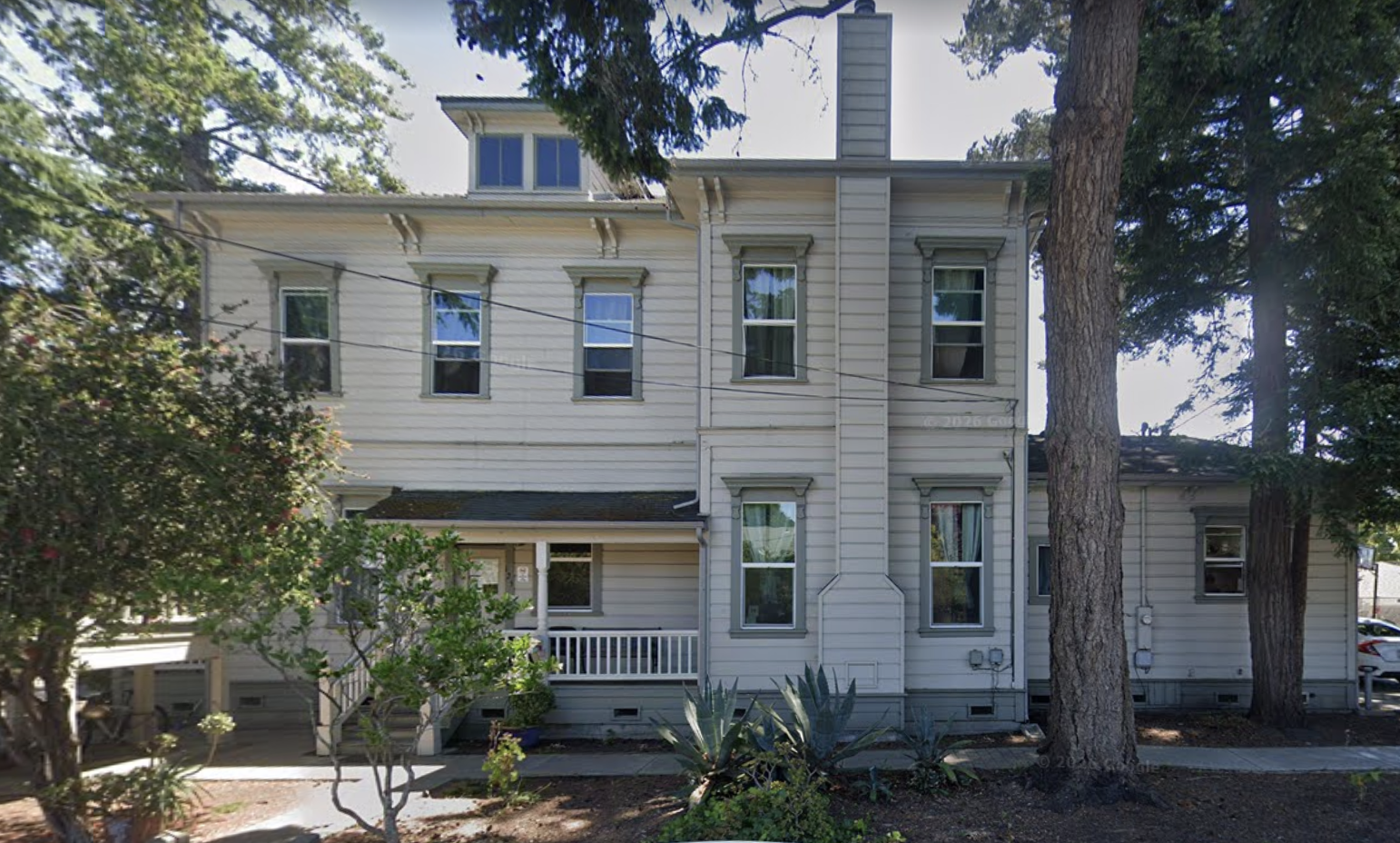 Two-story white residential building with porch and tall trees